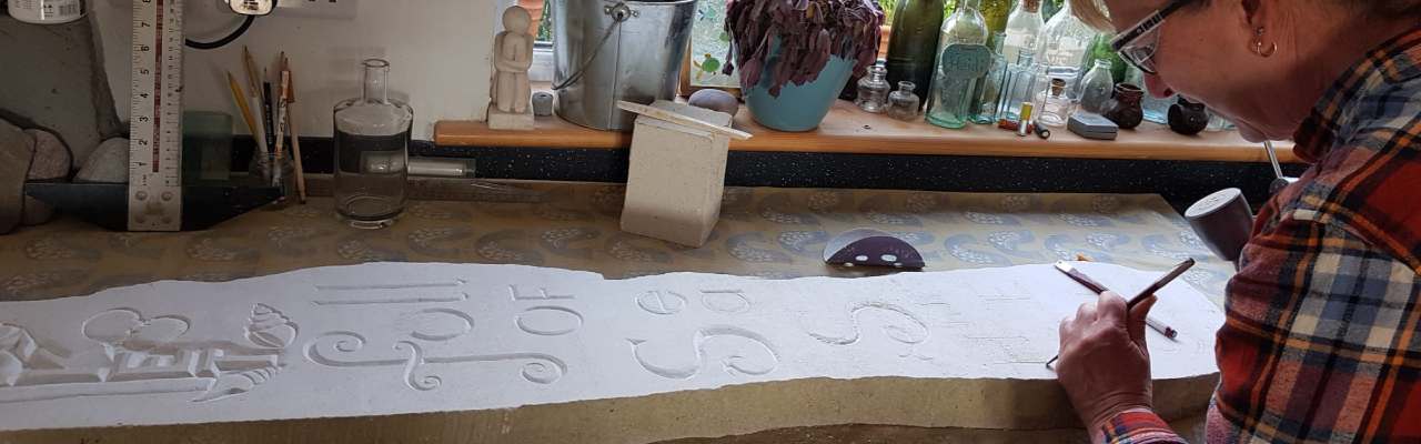 Image shows Heidi Mabbott hand carving a large piece of Portland stone with the script 'A POCKET full OF Sea SHhhELLs'with mallet and chisels.